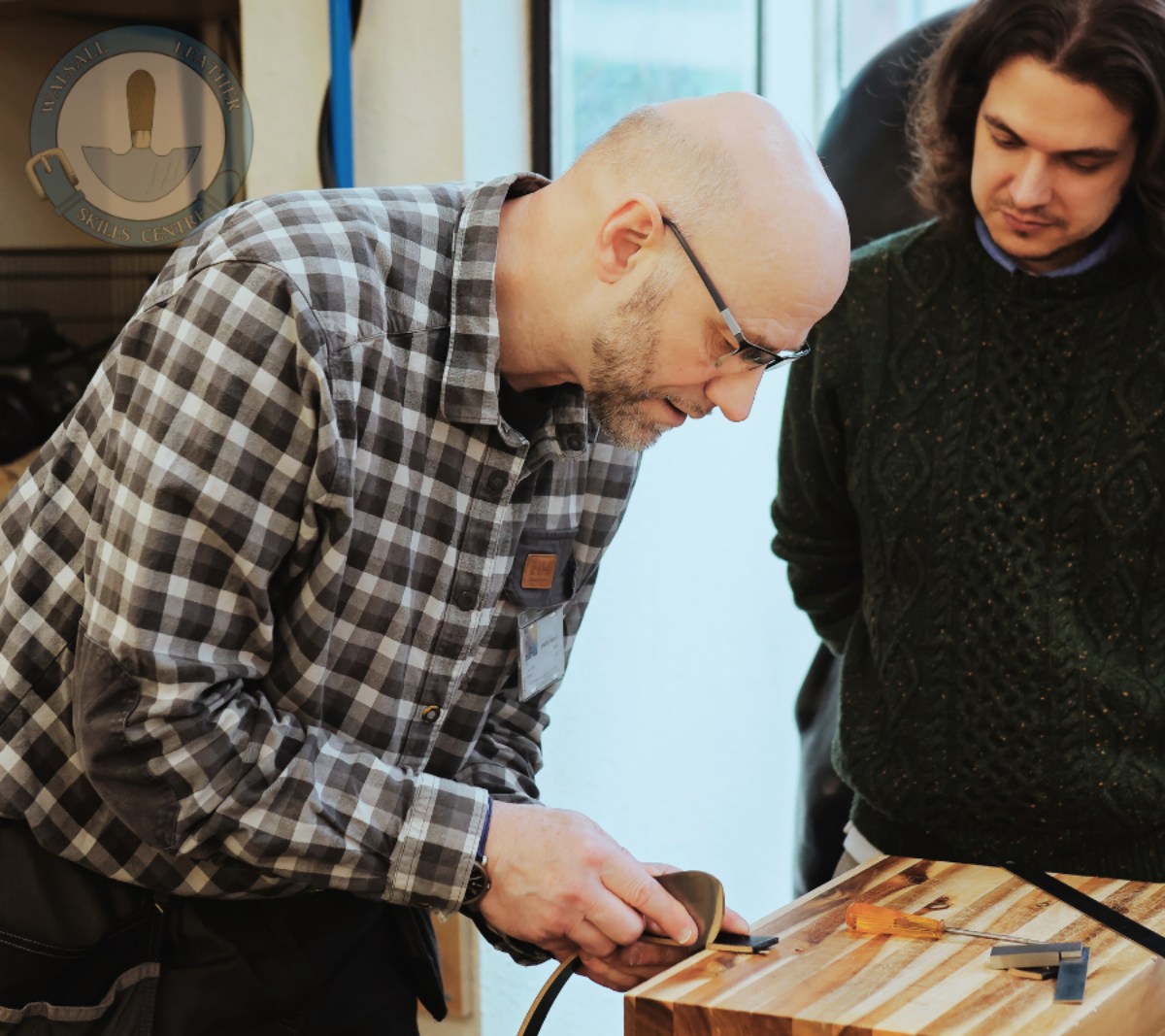 A photo from the Foundation Course at the Walsall Leather Skills Centre where the tutor is showing a student how to work properly with leather. There is a cutting board with some small tools and the tutor is burnishing the cut edges for a professional finish.