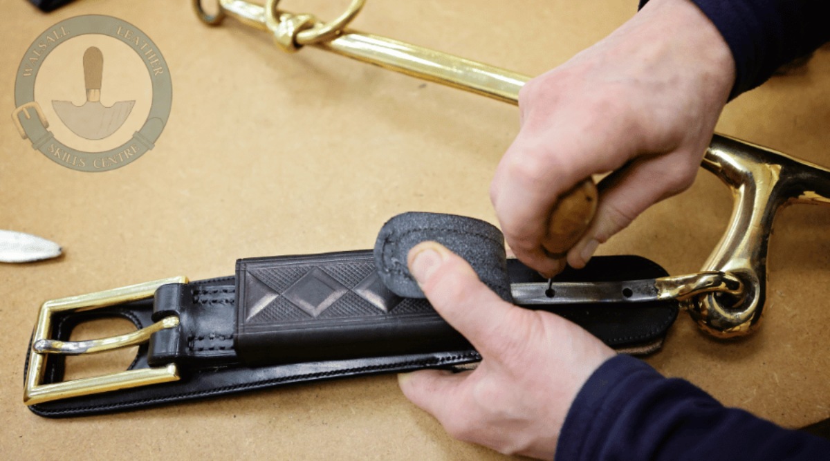 An image from the Advanced Harness Course at Walsall Leather Skills Centre or a student attaching harness hardware to a heavy duty piece of harness. The leather strap and buckle shows a decorated boxloop and the student is using an awl. There is WLSC Logo in the top left corner.